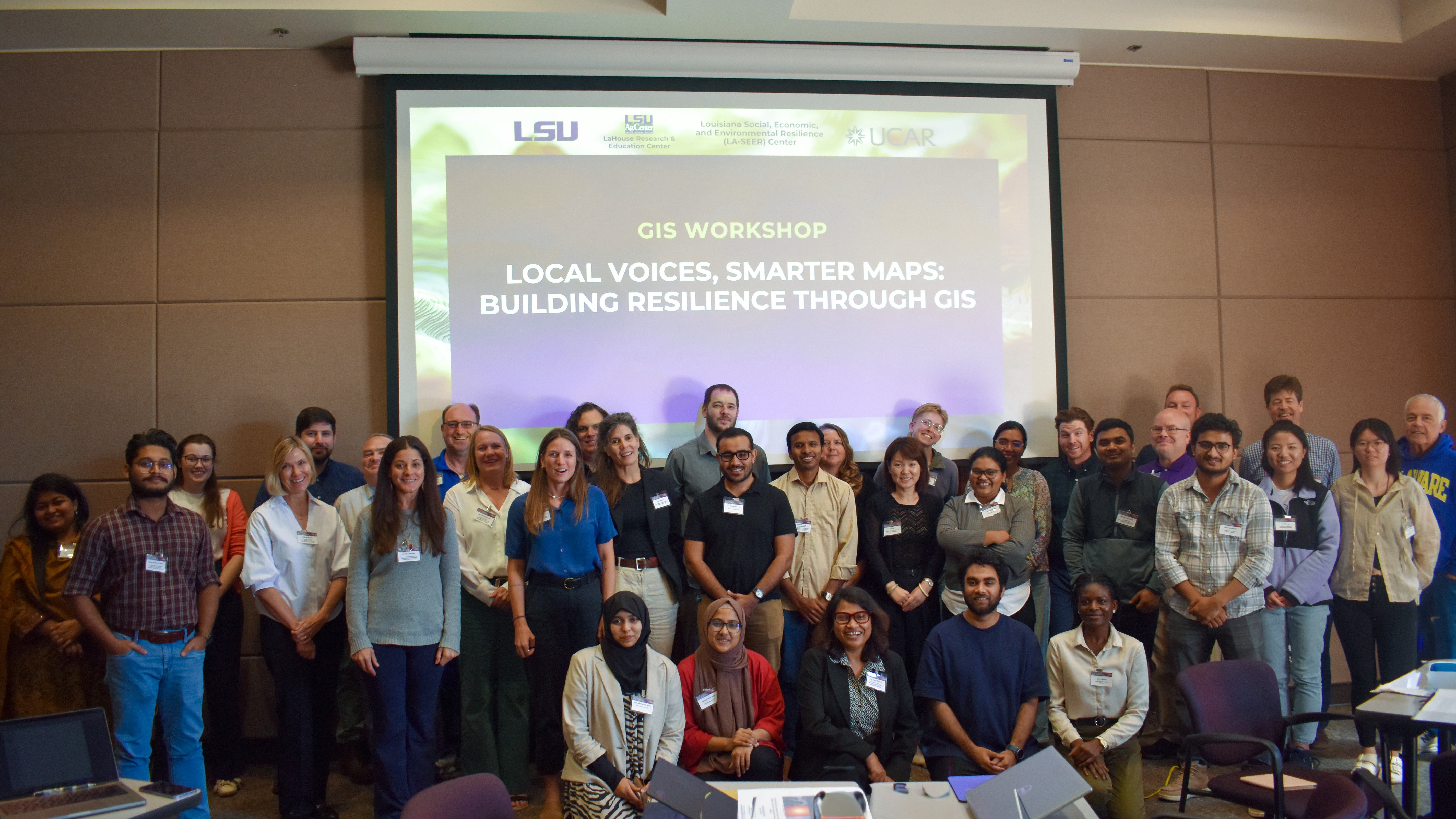 Attendees stand in front of a screen that says, "Local Voices Smarter Maps: Building resilience through GIS"