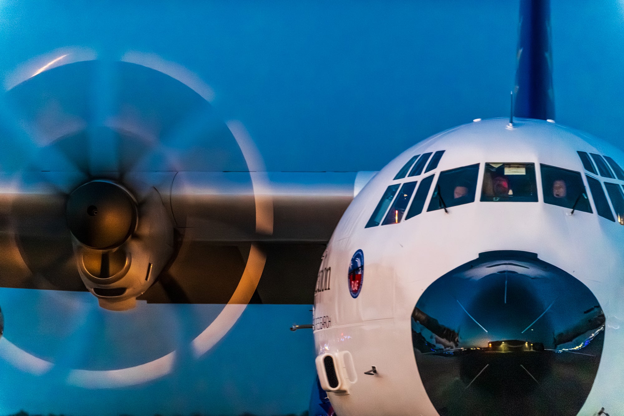 View of the front of a C-130 aircraft. A propeller is spinning and the pilot and co-pilot can be seen through the front windows.