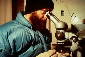 Charlie wearing a jacket and hat in the cold lab, studying ice under microscope.