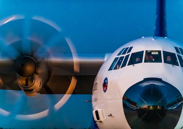 View of the front of a C-130 aircraft. A propeller is spinning and the pilot and co-pilot can be seen through the front windows.