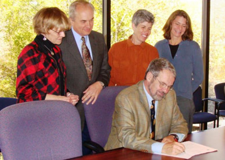 Photograph of Richard Anthes, sitting at desk, signing a document, other people